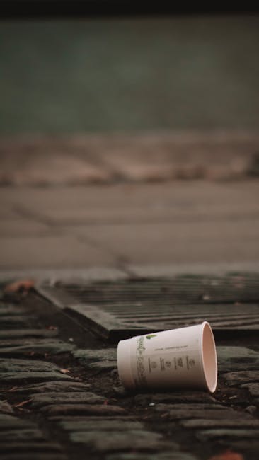 A white paper cup with printed text and logo lies on its side on a section of weathered cobblestone pavement near a metal drainage grate. The cup appears to be discarded, with its open top facing upwa